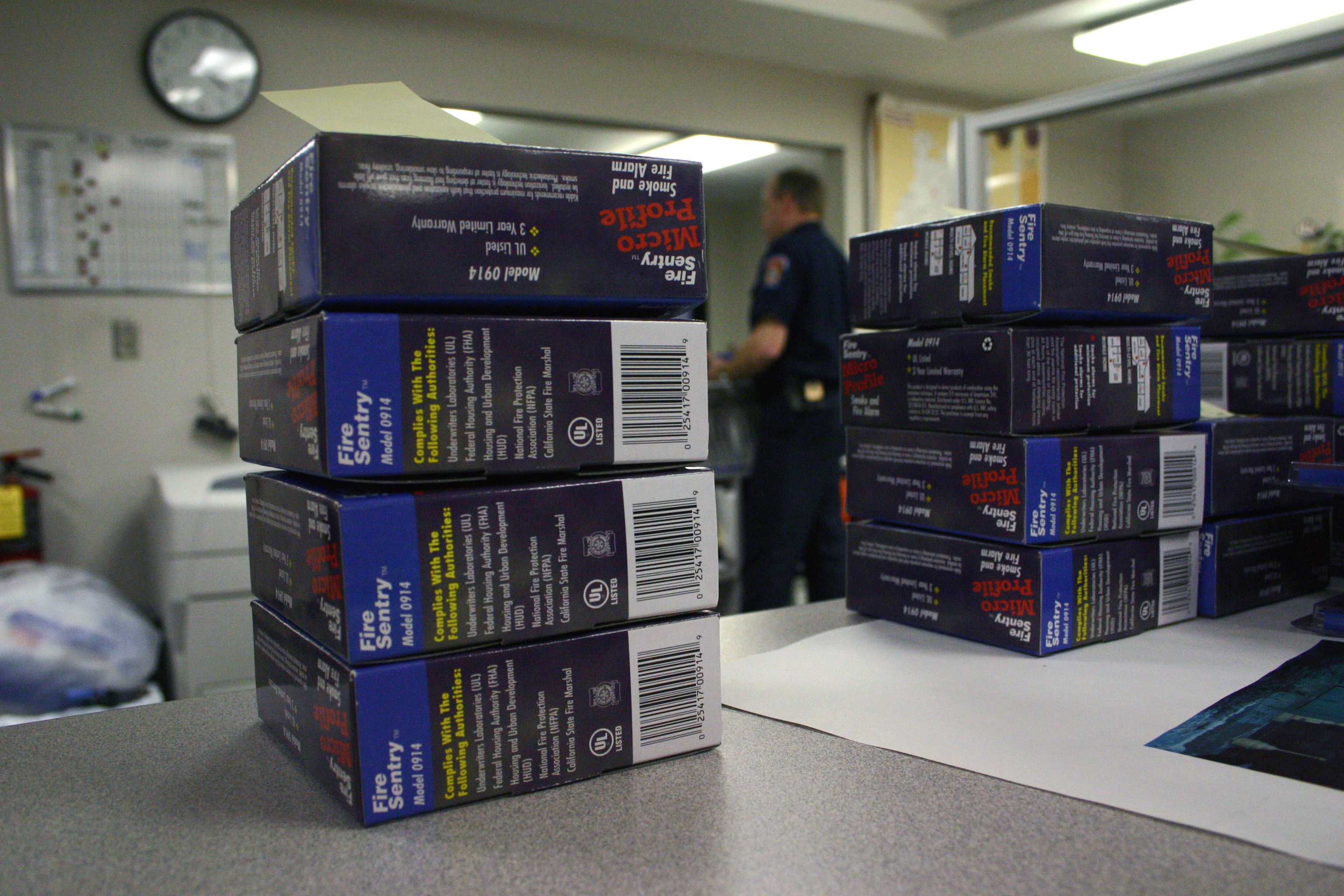 A stack of smoke alarm packages in the foreground with a uniformed officer standing in the background inside an office setting.