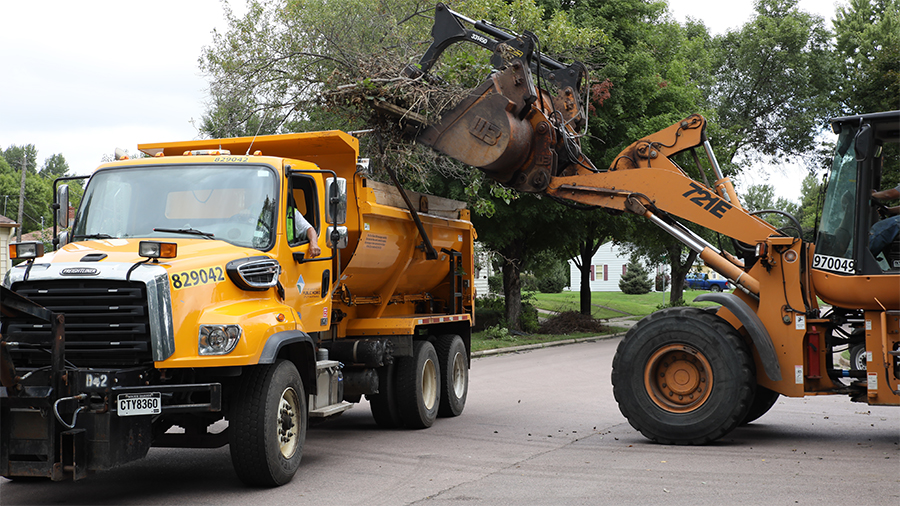 A yellow dump truck and a yellow telescopic handler working together to clear a large tree limb from a residential street. The telescopic handler is lifting the limb into the dump truck's open bed.